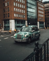 A green taxi is driving on an urban street surrounded by tall buildings with a modern design. The taxi has distinctive branding with the words 'Let's go!' visible on the side. The street is relatively empty with minimal traffic signs and road markings.