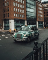 A green taxi is driving on an urban street surrounded by tall buildings with a modern design. The taxi has distinctive branding with the words 'Let's go!' visible on the side. The street is relatively empty with minimal traffic signs and road markings.