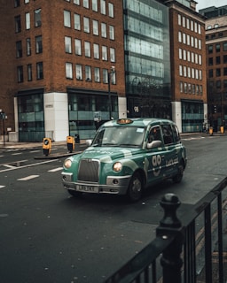 A green taxi is driving on an urban street surrounded by tall buildings with a modern design. The taxi has distinctive branding with the words 'Let's go!' visible on the side. The street is relatively empty with minimal traffic signs and road markings.