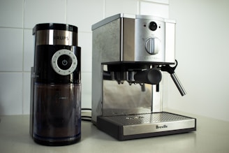 On a kitchen counter, a coffee grinder and an espresso machine are placed side by side. The coffee grinder, branded 'Krups', has a modern design with a black finish and a clear container showing coffee grounds inside. The adjacent espresso machine from 'Breville' has a stainless steel finish with a steam wand on the right side.