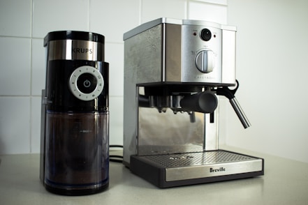 On a kitchen counter, a coffee grinder and an espresso machine are placed side by side. The coffee grinder, branded 'Krups', has a modern design with a black finish and a clear container showing coffee grounds inside. The adjacent espresso machine from 'Breville' has a stainless steel finish with a steam wand on the right side.
