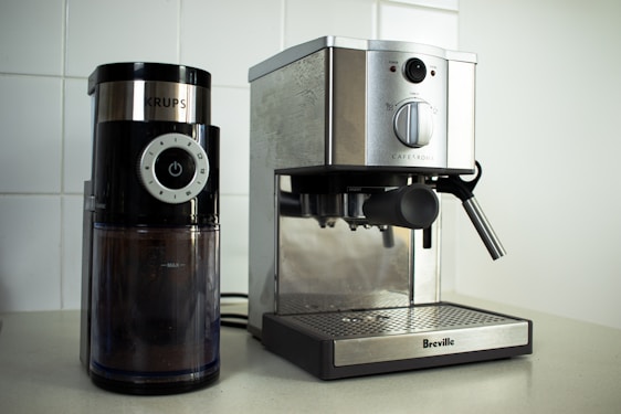 On a kitchen counter, a coffee grinder and an espresso machine are placed side by side. The coffee grinder, branded 'Krups', has a modern design with a black finish and a clear container showing coffee grounds inside. The adjacent espresso machine from 'Breville' has a stainless steel finish with a steam wand on the right side.