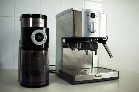On a kitchen counter, a coffee grinder and an espresso machine are placed side by side. The coffee grinder, branded 'Krups', has a modern design with a black finish and a clear container showing coffee grounds inside. The adjacent espresso machine from 'Breville' has a stainless steel finish with a steam wand on the right side.