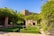 A beautiful garden scene with neatly trimmed hedges arranged in geometric patterns. Stone archways covered in lush green vines and surrounded by well-maintained plants frame the background. The sky is clear and blue, adding a serene atmosphere to the setting.