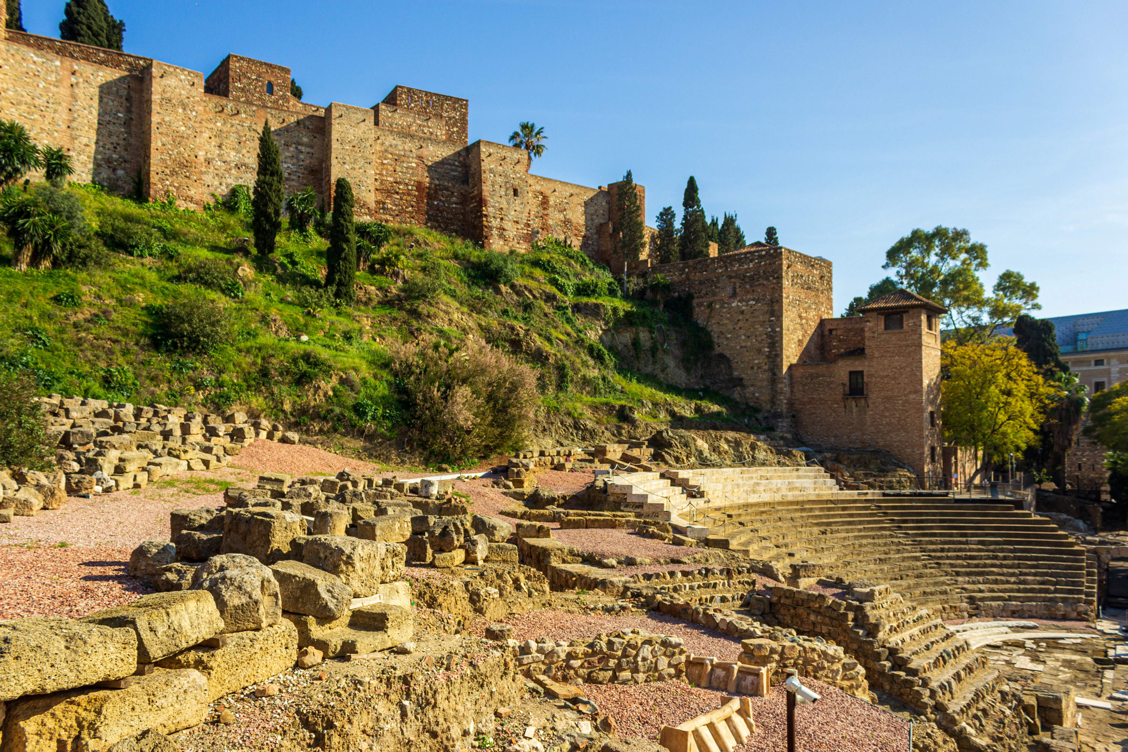 The excavated stone tiers of Malaga's Roman Theatre in the foreground, with the honey-coloured battlements of the Alcazaba fortress rising on the green hillside above under a clear blue sky.