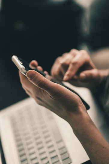 A person using a smartphone above a laptop keyboard in a workspace