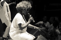 A performer with voluminous hair is kneeling and passionately singing into a microphone on stage. The audience is close, with several people visible in the low light, some holding up their phones to capture the moment. The scene is in black and white, giving it a classic and timeless feel.