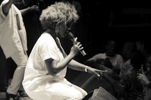 A performer with voluminous hair is kneeling and passionately singing into a microphone on stage. The audience is close, with several people visible in the low light, some holding up their phones to capture the moment. The scene is in black and white, giving it a classic and timeless feel.