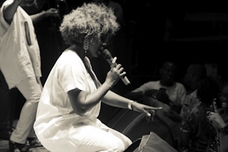 A performer with voluminous hair is kneeling and passionately singing into a microphone on stage. The audience is close, with several people visible in the low light, some holding up their phones to capture the moment. The scene is in black and white, giving it a classic and timeless feel.