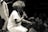 A performer with voluminous hair is kneeling and passionately singing into a microphone on stage. The audience is close, with several people visible in the low light, some holding up their phones to capture the moment. The scene is in black and white, giving it a classic and timeless feel.