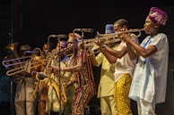 A vibrant group of musicians wearing colorful traditional clothing plays various brass instruments. The scene is lively with a sense of rhythm and energy as the musicians focus on their performance, suggesting a live music event. The backdrop is dark, highlighting the performers under stage lighting.