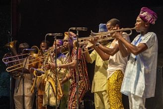 A vibrant group of musicians wearing colorful traditional clothing plays various brass instruments. The scene is lively with a sense of rhythm and energy as the musicians focus on their performance, suggesting a live music event. The backdrop is dark, highlighting the performers under stage lighting.