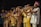 A vibrant group of musicians wearing colorful traditional clothing plays various brass instruments. The scene is lively with a sense of rhythm and energy as the musicians focus on their performance, suggesting a live music event. The backdrop is dark, highlighting the performers under stage lighting.