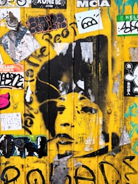 A vibrant yellow wall adorned with numerous stickers and graffiti. Among the graffiti, there is a large, detailed stencil of a person's face. Surrounding this are various colorful stickers and tags, including black-and-white designs and multicolored artwork.