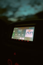 A car dashboard displaying a digital screen with music playing. The screen shows a song by an artist, with album art clearly visible. The surrounding dashboard is dimly lit, and the ambient light outside the car is low, giving a nighttime feel.