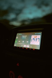 A car dashboard displaying a digital screen with music playing. The screen shows a song by an artist, with album art clearly visible. The surrounding dashboard is dimly lit, and the ambient light outside the car is low, giving a nighttime feel.