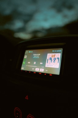 A car dashboard displaying a digital screen with music playing. The screen shows a song by an artist, with album art clearly visible. The surrounding dashboard is dimly lit, and the ambient light outside the car is low, giving a nighttime feel.