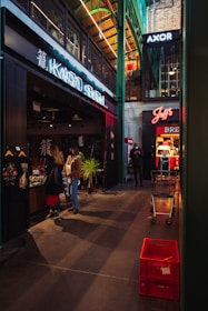 A modern indoor market with various stores and signage, including a sushi restaurant and other shops. People are walking and browsing near the stores. There are shopping carts and red crates visible on the floor, and the architecture features high ceilings with exposed beams.