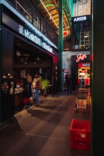 A modern indoor market with various stores and signage, including a sushi restaurant and other shops. People are walking and browsing near the stores. There are shopping carts and red crates visible on the floor, and the architecture features high ceilings with exposed beams.