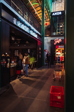 A modern indoor market with various stores and signage, including a sushi restaurant and other shops. People are walking and browsing near the stores. There are shopping carts and red crates visible on the floor, and the architecture features high ceilings with exposed beams.