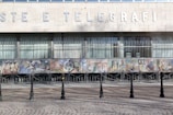 A building facade features large block letters spelling out 'POSTE E TELEGRAFI' above a series of tall windows with vertical blinds. Below the windows, a colorful mosaic mural depicts various abstract and figurative elements, including geometric shapes and figures in diverse settings. The foreground consists of a cobblestone pavement with a line of black bollards connected by chains.