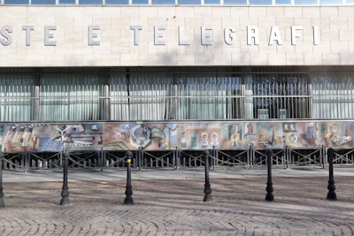A building facade features large block letters spelling out 'POSTE E TELEGRAFI' above a series of tall windows with vertical blinds. Below the windows, a colorful mosaic mural depicts various abstract and figurative elements, including geometric shapes and figures in diverse settings. The foreground consists of a cobblestone pavement with a line of black bollards connected by chains.
