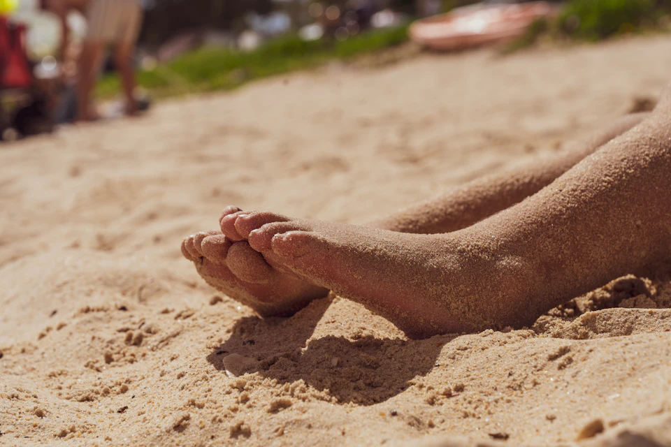 Close-up view of a person's feet resting on sandy beach with scattered rocks nearby