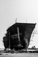 A large, unfinished ship is resting on a dry dock with metal scaffolding protruding from its sides. The ship has visible ladders and metal support beams. In the background, there's another ship on the water. The scene is set in an industrial area with sparse vegetation on the ground and a clear sky overhead.