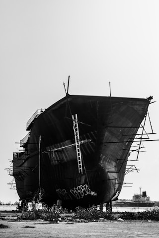 A large, unfinished ship is resting on a dry dock with metal scaffolding protruding from its sides. The ship has visible ladders and metal support beams. In the background, there's another ship on the water. The scene is set in an industrial area with sparse vegetation on the ground and a clear sky overhead.