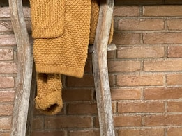 A cozy knitted mustard yellow sweater draped over a rustic wooden ladder against a brick wall.