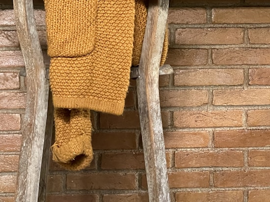 A cozy knitted mustard yellow sweater draped over a rustic wooden ladder against a brick wall.