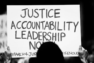 A person is holding a protest sign with bold text reading 'JUSTICE ACCOUNTABILITY LEADERSHIP NOW' written on it. The sign also includes smaller text at the bottom stating 'MARCH4JUSTICE' and '#ENOUGHISENOUGH'. The image is in black and white, enhancing a serious and impactful tone.