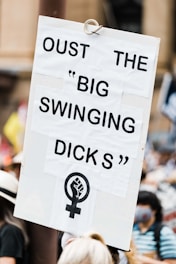 A protest sign with the text 'OUST THE 'BIG SWINGING DICKS'' and a feminist symbol with a raised fist held in a crowd. People in the background are partially visible, and some appear to be wearing masks.