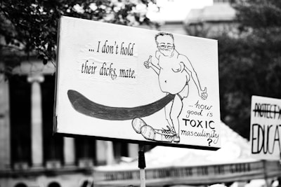 A protest sign with a cartoon drawing of a man holding his hands up, accompanied by the text questioning toxic masculinity. The illustration includes exaggerated features and humorous intent. Other protest signs with text are visible in the background.