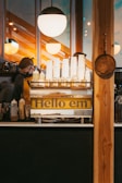 A coffee shop interior featuring a counter with stacked white cups on a metal tray labeled 'Hello em'. Behind the counter, two people are partially visible, preparing beverages. The warm glow from hanging lights and wooden textures create a cozy ambiance. A pan is hanging on the wooden panel at the right side of the image.