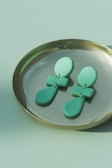 A pair of green geometric earrings rests on a brass dish. The earrings feature oval shapes on top connected to rectangular segments, leading to teardrop-shaped designs at the bottom. The colors vary between light and dark green, and the dish sits on a matching green surface.