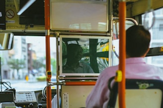The interior of a public bus with a focus on the driver's area. The rear view mirror prominently displays a reflection of the driver, while the perspective suggests the viewer is seated behind the driver. Various equipment and signage typical of buses are visible, creating an urban transport atmosphere.
