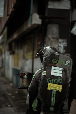 A motorcyclist is wearing a helmet and a jacket with the 'Grab' logo, sitting on a motorcycle in an urban setting. The background features blurred buildings, suggesting a city alley or street with graffiti and worn structures.