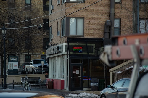 A brick building with a storefront displaying a sign for a coffee shop. Two bicycles are parked on the sidewalk in front of metal chairs and a small table. A pickup truck is visible, parked near the building. Bare trees and overhead wires are also present, along with a street sign.