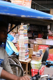 A street market stall is filled with stacks of books and various office supplies. Several large piles of books are tied together, surrounded by packages of paper and some cardboard boxes. A sign with Hindi text and other advertisements is visible above. In the foreground, a part of a bicycle or rickshaw is visible with a person partially in view.
