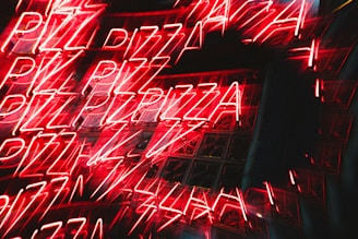 Bright neon signs of the word 'PIZZA' are repeated multiple times, creating a dynamic and energetic visual effect with reflections on a glass surface.