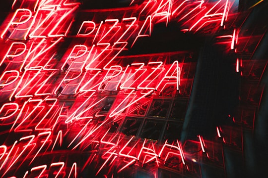 Bright neon signs of the word 'PIZZA' are repeated multiple times, creating a dynamic and energetic visual effect with reflections on a glass surface.
