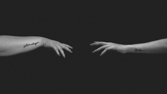 Two hands with tattoos on their forearms reach towards each other against a dark background, creating a sense of connection or longing.