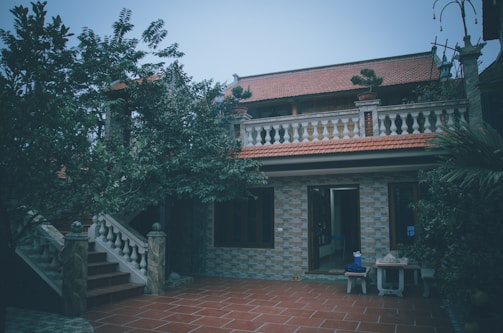 A two-story house with a red tiled roof and brick walls is surrounded by lush greenery. The front features a tiled courtyard with stairs leading to an upper balcony. Several large trees frame the building, casting shadows in the area.