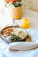 A stylishly arranged breakfast setting includes a bowl with a variety of food items, featuring beans, grains, and leafy greens. A cozy napkin is tucked under the bowl on a white tablecloth. Next to the bowl, a glass of orange juice adds a splash of vibrant color. In the background, a wicker basket with flowers enhances the overall aesthetic, contributing to a warm and inviting atmosphere.
