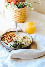 A stylishly arranged breakfast setting includes a bowl with a variety of food items, featuring beans, grains, and leafy greens. A cozy napkin is tucked under the bowl on a white tablecloth. Next to the bowl, a glass of orange juice adds a splash of vibrant color. In the background, a wicker basket with flowers enhances the overall aesthetic, contributing to a warm and inviting atmosphere.