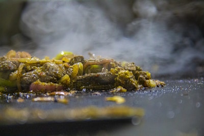 A close-up view of sizzling, seasoned meat mixed with onions and possibly green peppers on a hot surface. The scene is enveloped in rising steam, giving an impression of freshly cooked food.