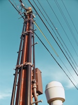 A tall utility pole with multiple wires and cables attached, reaching up against the backdrop of a clear blue sky. The pole appears to be constructed from metal and has a rust color, while the cables are dark and extend horizontally. Near the base of the pole, there is a white cylindrical object.