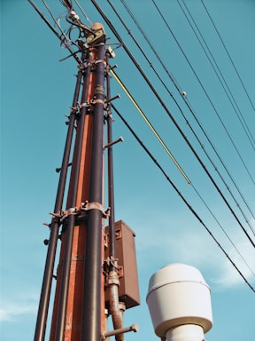 A tall utility pole with multiple wires and cables attached, reaching up against the backdrop of a clear blue sky. The pole appears to be constructed from metal and has a rust color, while the cables are dark and extend horizontally. Near the base of the pole, there is a white cylindrical object.