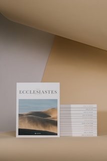 A modern, minimalist arrangement featuring a prominent book titled 'Ecclesiastes' with an image of sand dunes on its cover. Next to it, a neatly stacked collection of similar books with various titles, including 'Book of Genesis' and 'Book of Job'. The background displays a smooth gradient of beige and tan colors.